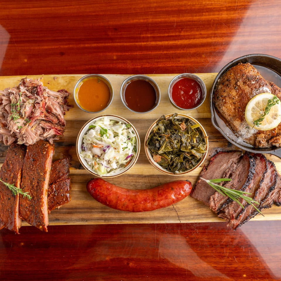 Overhead barbecue spread with ribs, pulled pork, sausage, sliced brisket, roasted chicken, sauces, coleslaw, and greens on wooden boards.