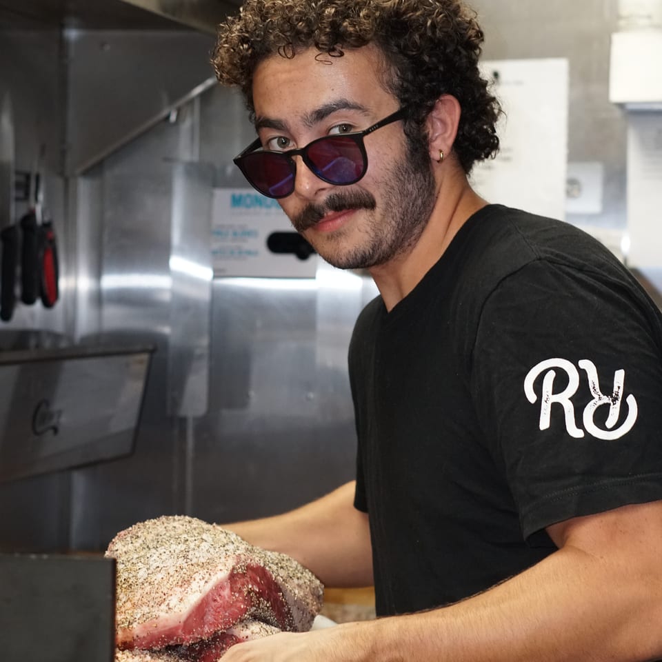 Restaurant worker wearing sunglasses slices a heavily seasoned raw beef roast on a cutting board in a commercial kitchen.