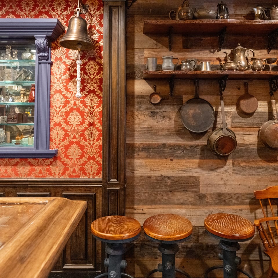 Rustic restaurant interior with wooden shelves displaying vintage cookware, red patterned wallpaper, a brass bell, stools, and a glass cabinet.