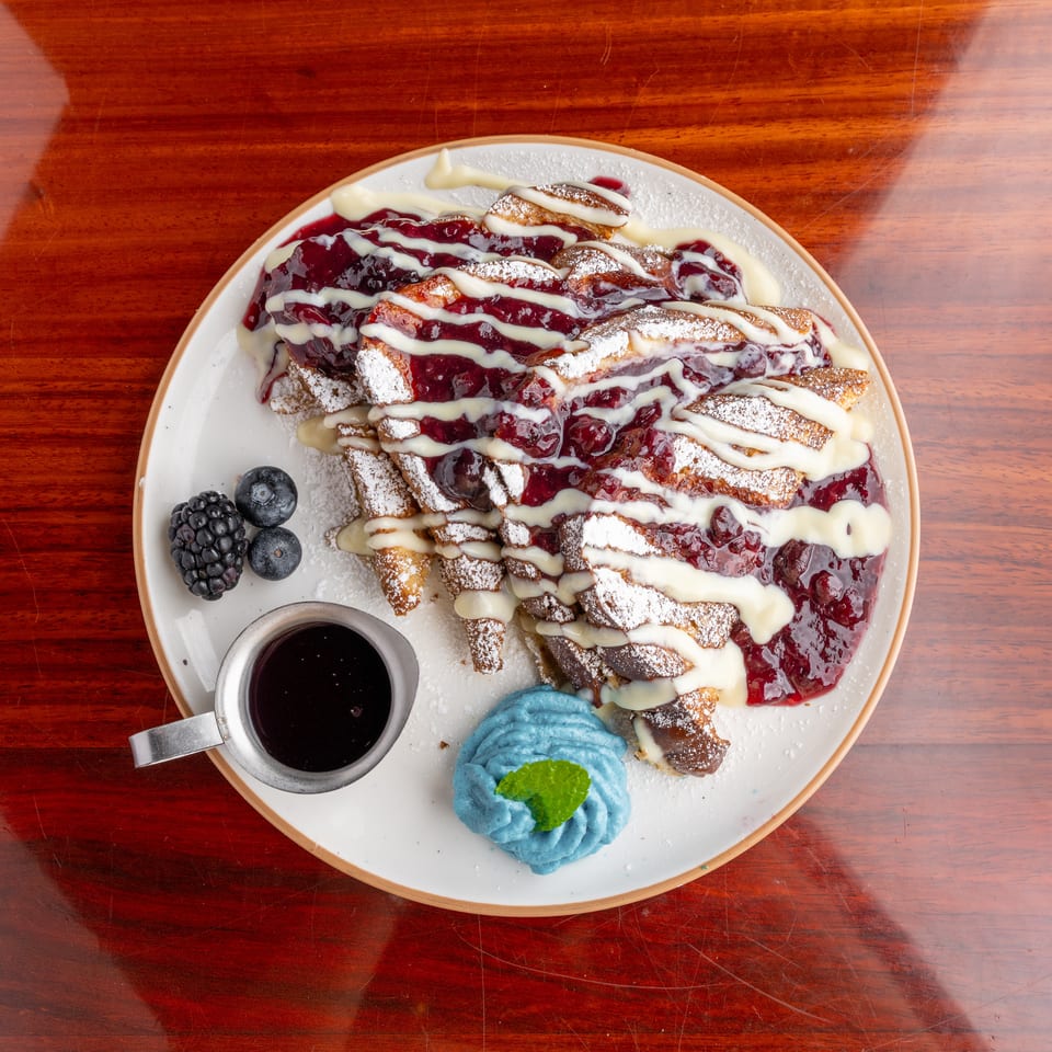 Overhead view of berry-topped French toast with powdered sugar, cream drizzle, berries, syrup, and blue whipped cream on a plate.