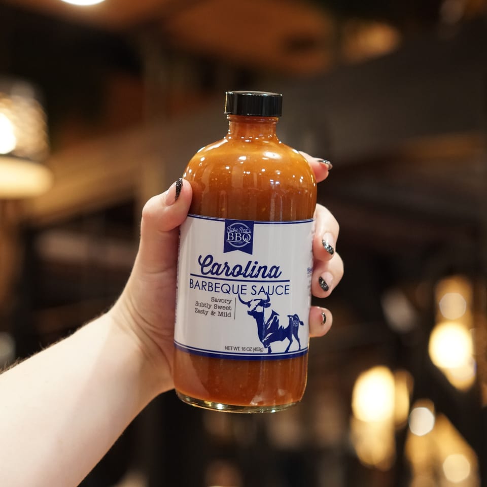 Hand holding a bottle of Ruby Red’s Carolina barbecue sauce against a warmly lit, blurred restaurant background.