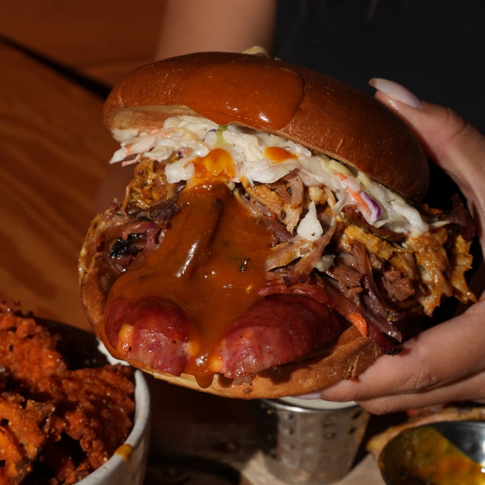 Close-up of a loaded barbecue sandwich with pulled meat, sausage, coleslaw, and barbecue sauce, held above a restaurant table.