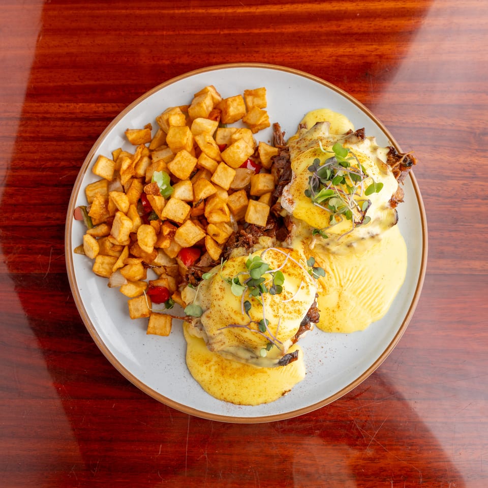 Plate of eggs Benedict topped with hollandaise and microgreens, served with crispy diced potatoes on a wooden table.