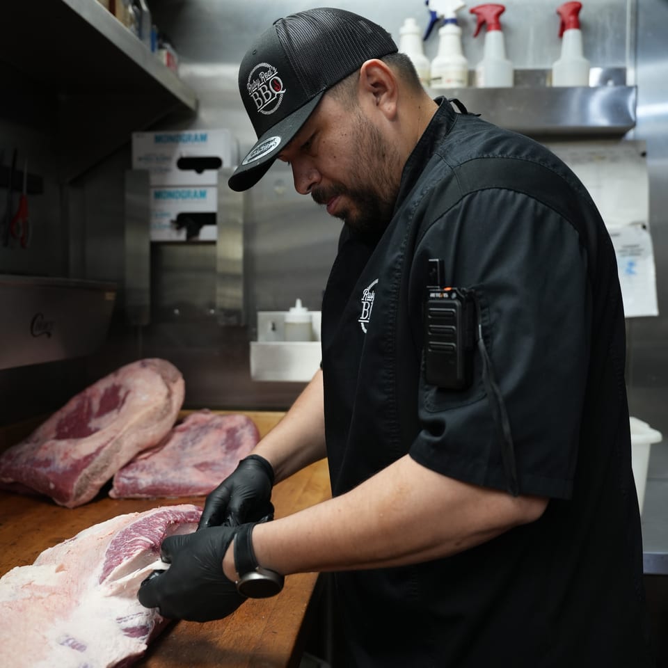 Barbecue chef in a commercial kitchen trims raw meat on a wooden cutting board while wearing gloves and branded black attire.