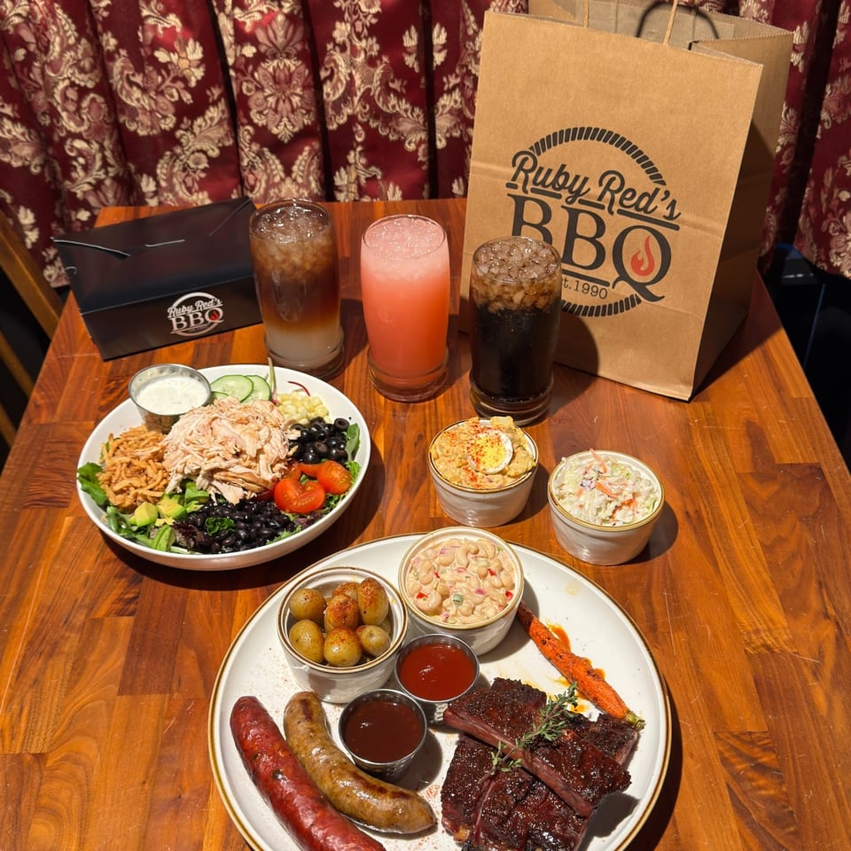 Ruby Red’s BBQ table spread with ribs, sausages, salads, sides, drinks, and branded bags against patterned curtains.