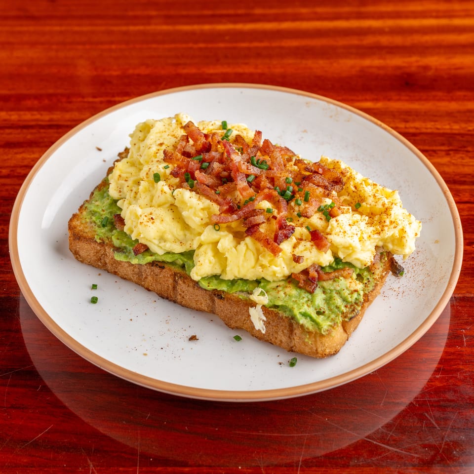 Avocado toast topped with scrambled eggs, crispy bacon, chives, and seasoning on a white plate.