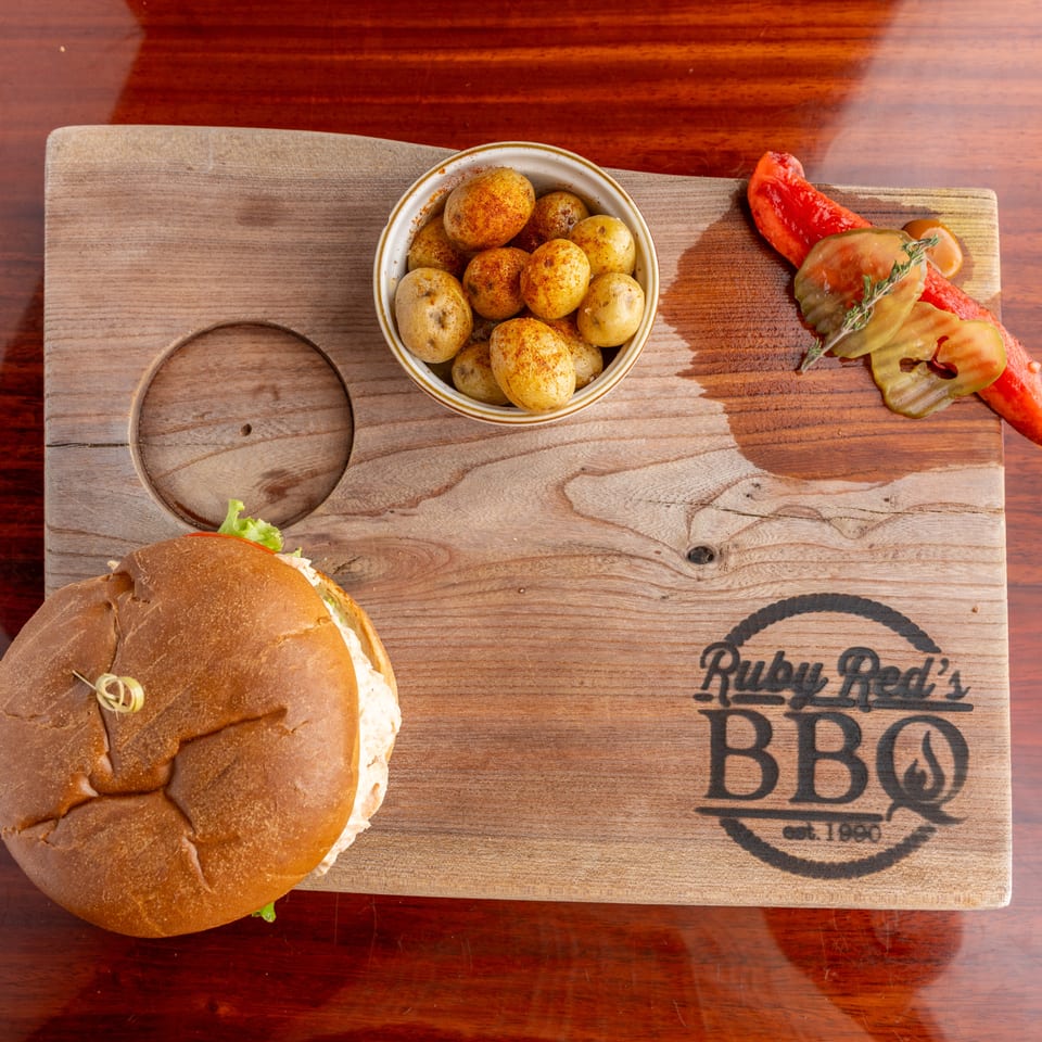 Burger, seasoned baby potatoes, and sliced tomatoes arranged on a branded wooden serving board.