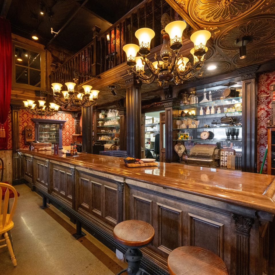 Ornate restaurant bar with dark wood paneling, red damask wallpaper, glowing chandeliers, mirrored shelves, and stools.