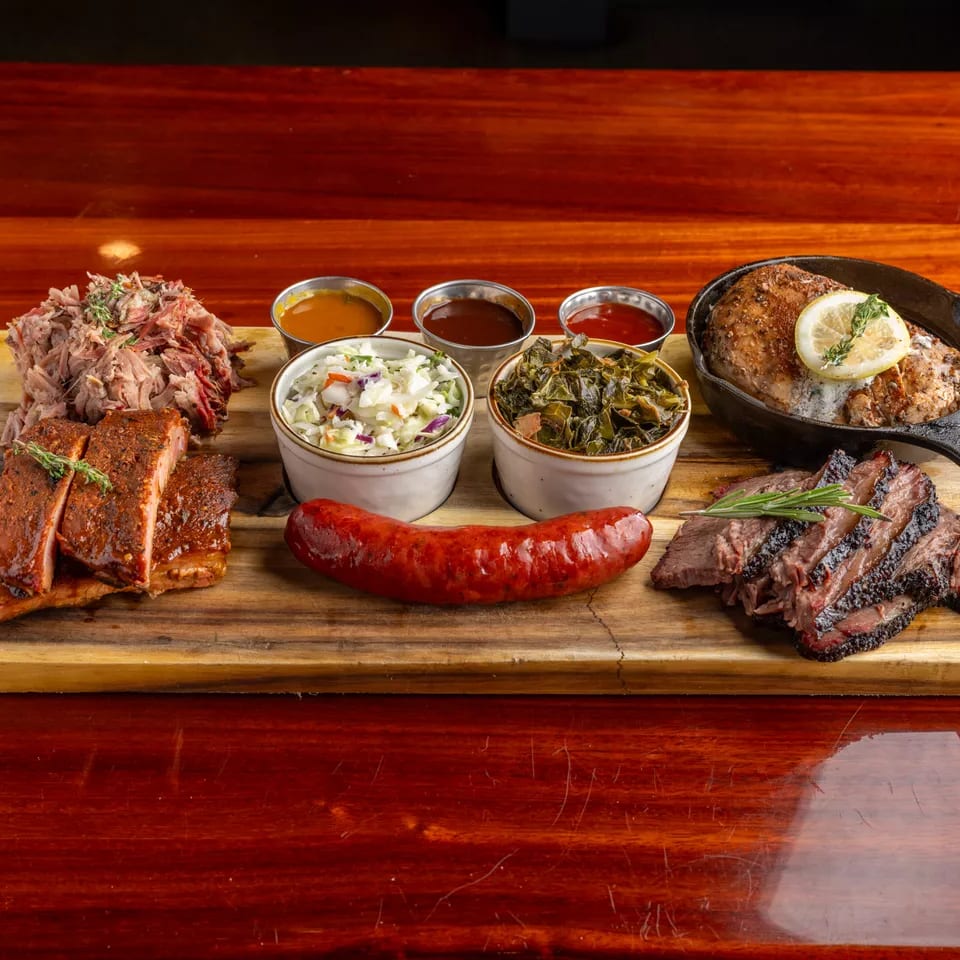 Barbecue platter on a wooden board with ribs, pulled pork, sausage, sliced brisket, chicken, sides, and dipping sauces.