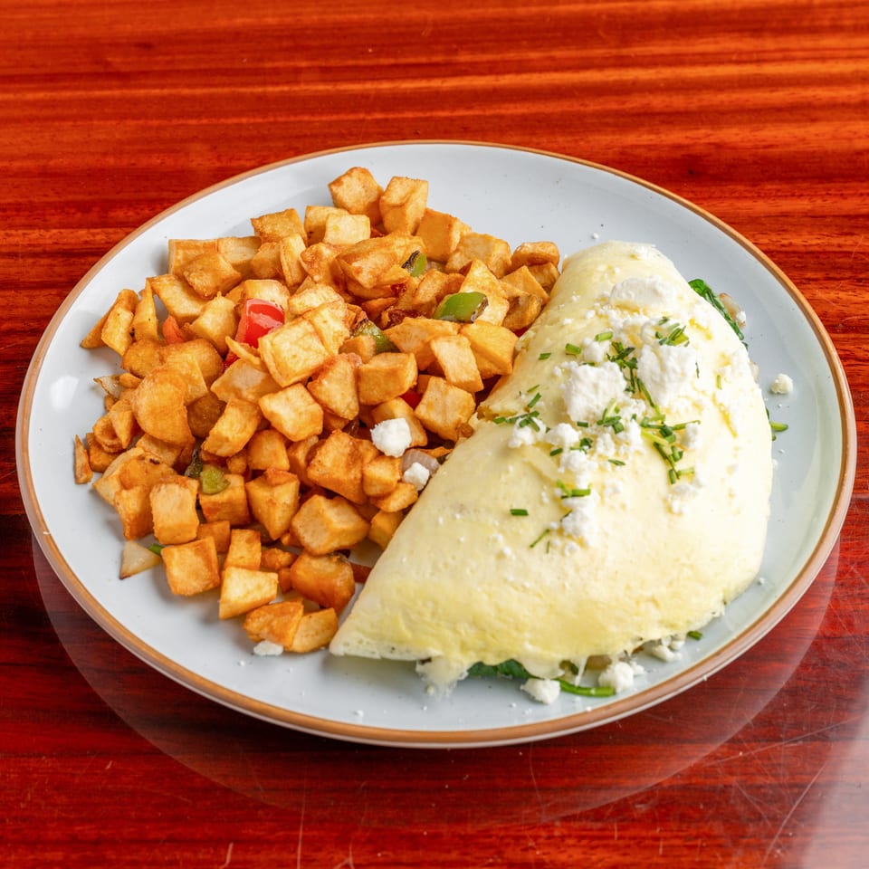 Cheese-filled omelet topped with chives and crumbled cheese, served alongside seasoned diced potatoes and vegetables on a plate.