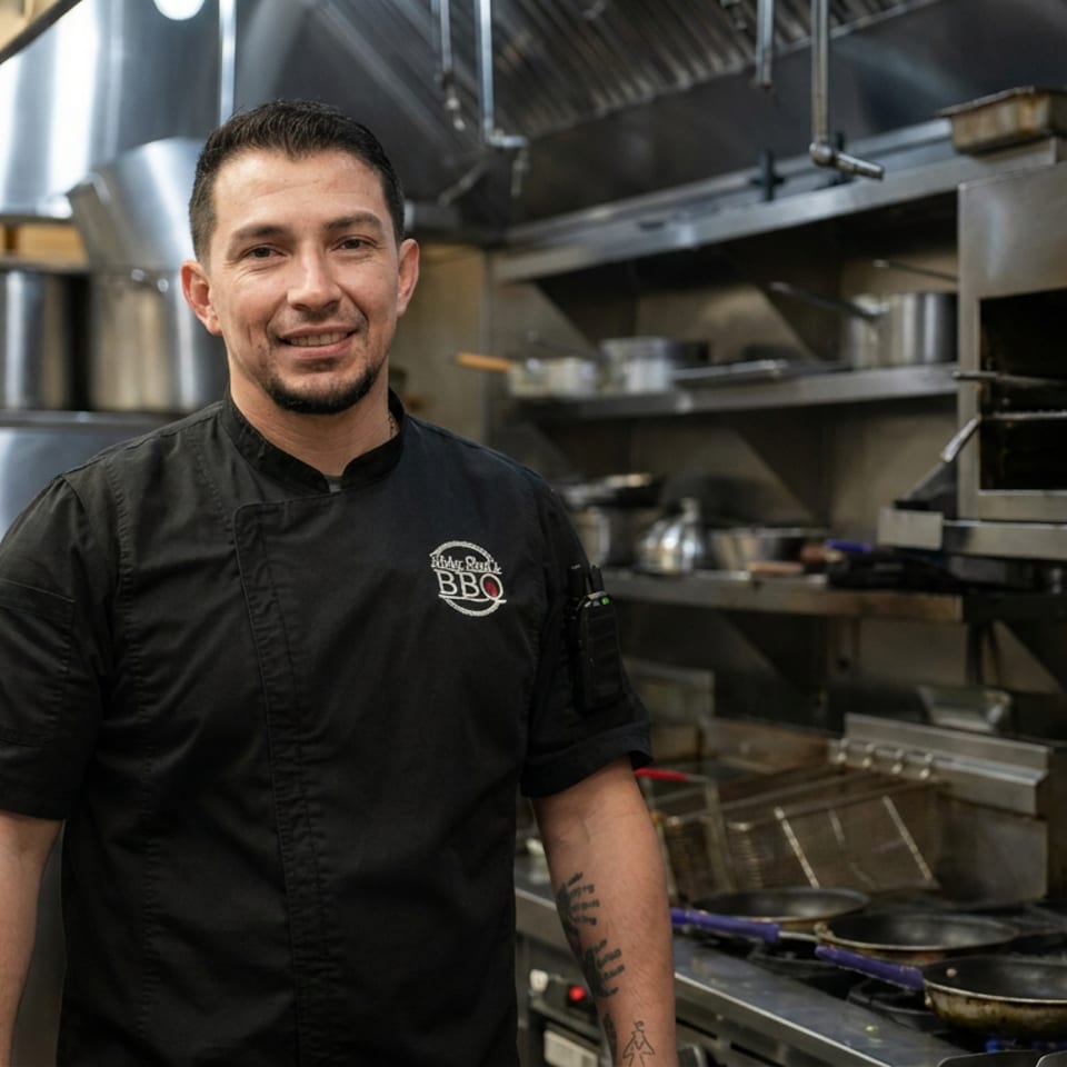 Smiling chef in a black barbecue-logo uniform stands inside a busy commercial restaurant kitchen beside ovens and stovetops.