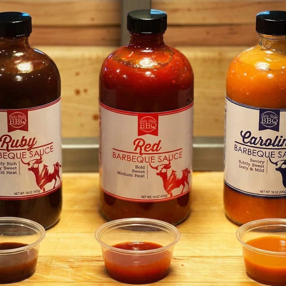 Three bottles of Ruby, Red, and Carolina barbecue sauces with matching tasting cups displayed on a wooden board.