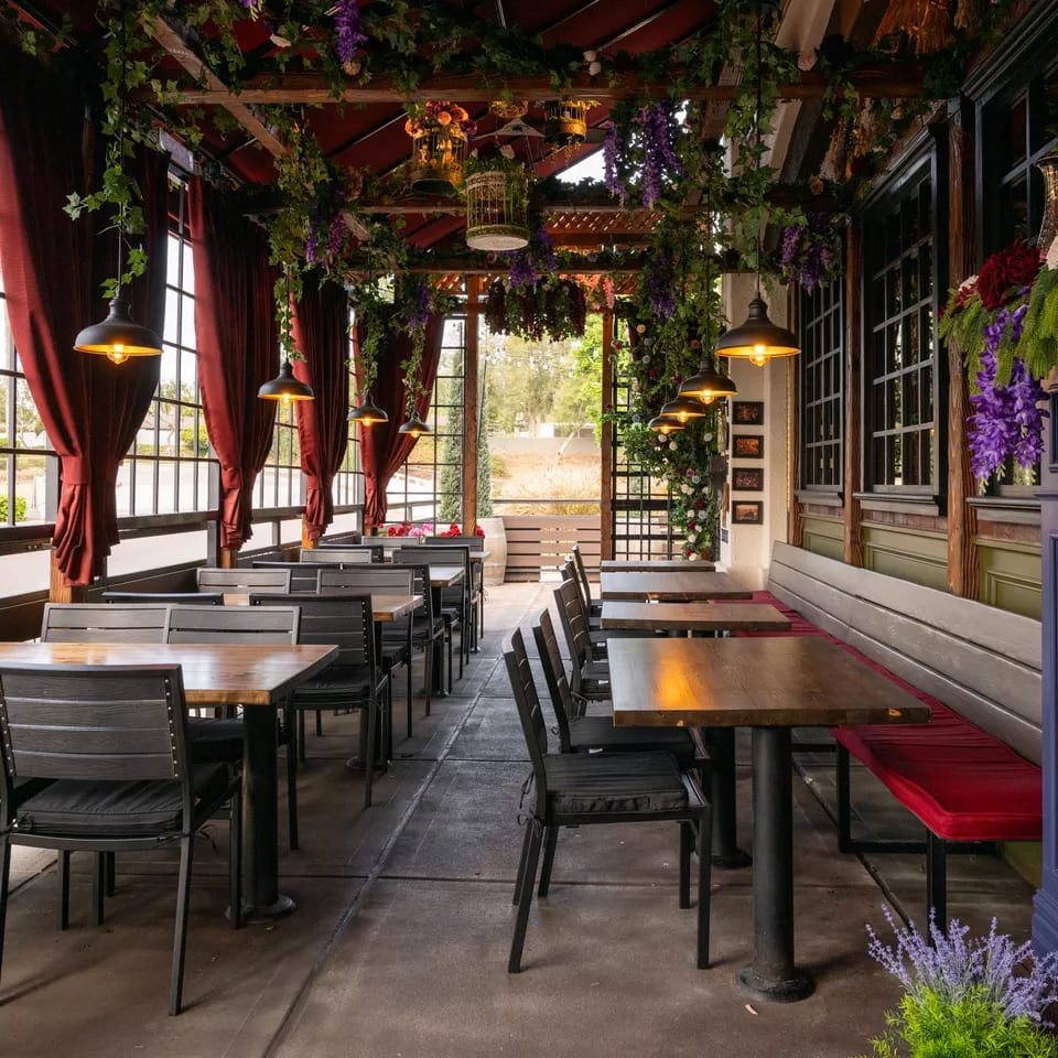 Elegant restaurant dining room with wooden tables, black chairs, red curtains, warm pendant lights, and abundant hanging greenery and flowers.