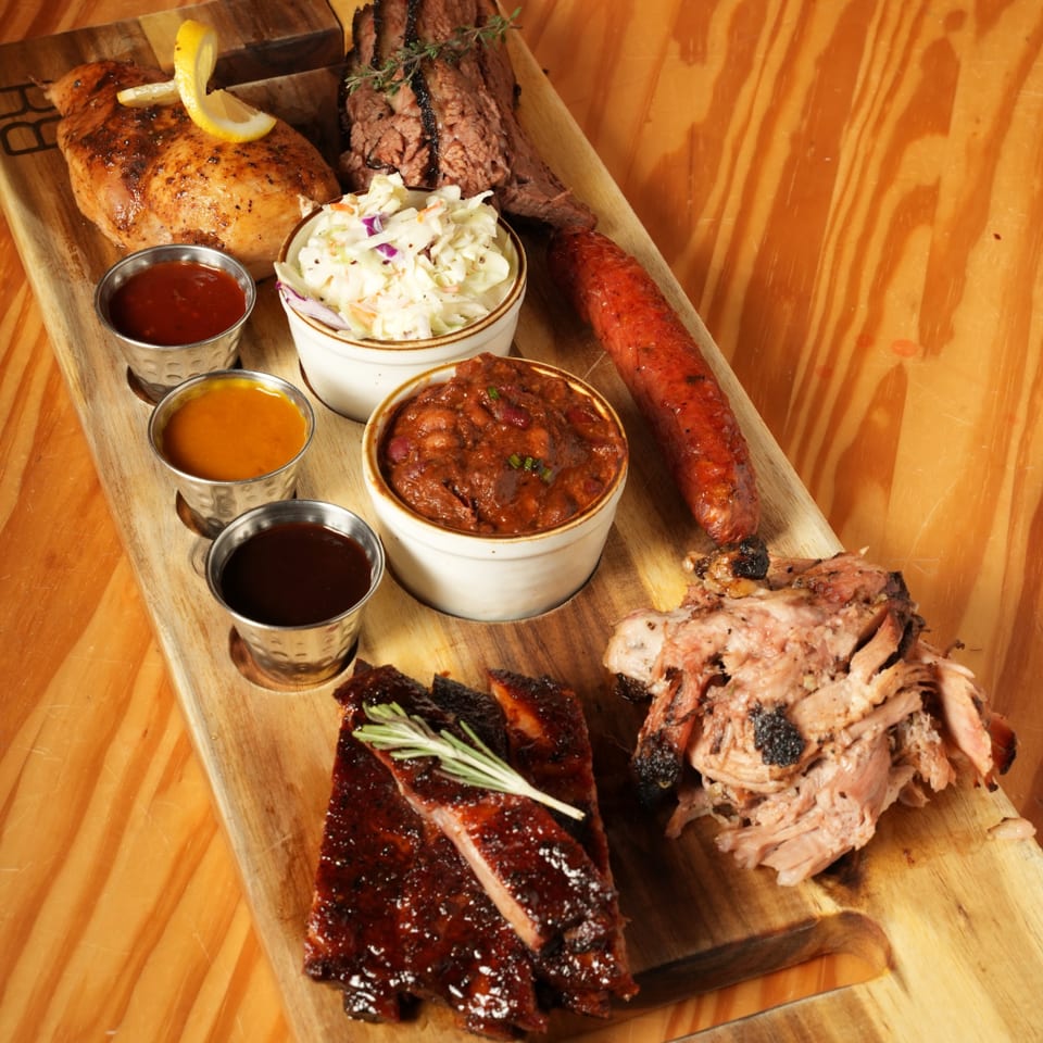 Barbecue platter on a wooden board with ribs, sliced steak, chicken, sausage, pulled pork, sides, and dipping sauces.