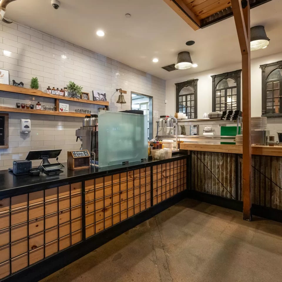 Rustic restaurant counter with wood paneling, tiled walls, shelves of bottles, coffee equipment, and industrial-style decor.