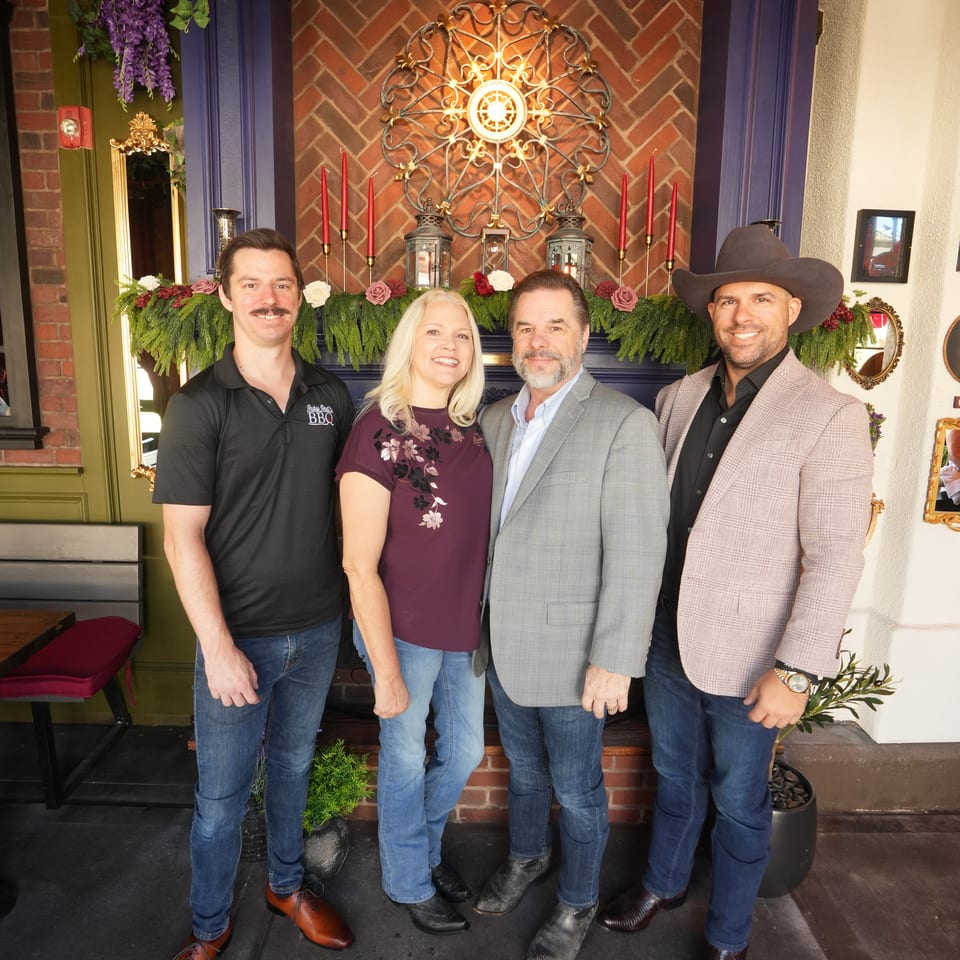 Four people pose in front of a decorated brick fireplace with floral garlands, candles, and ornate wall art.