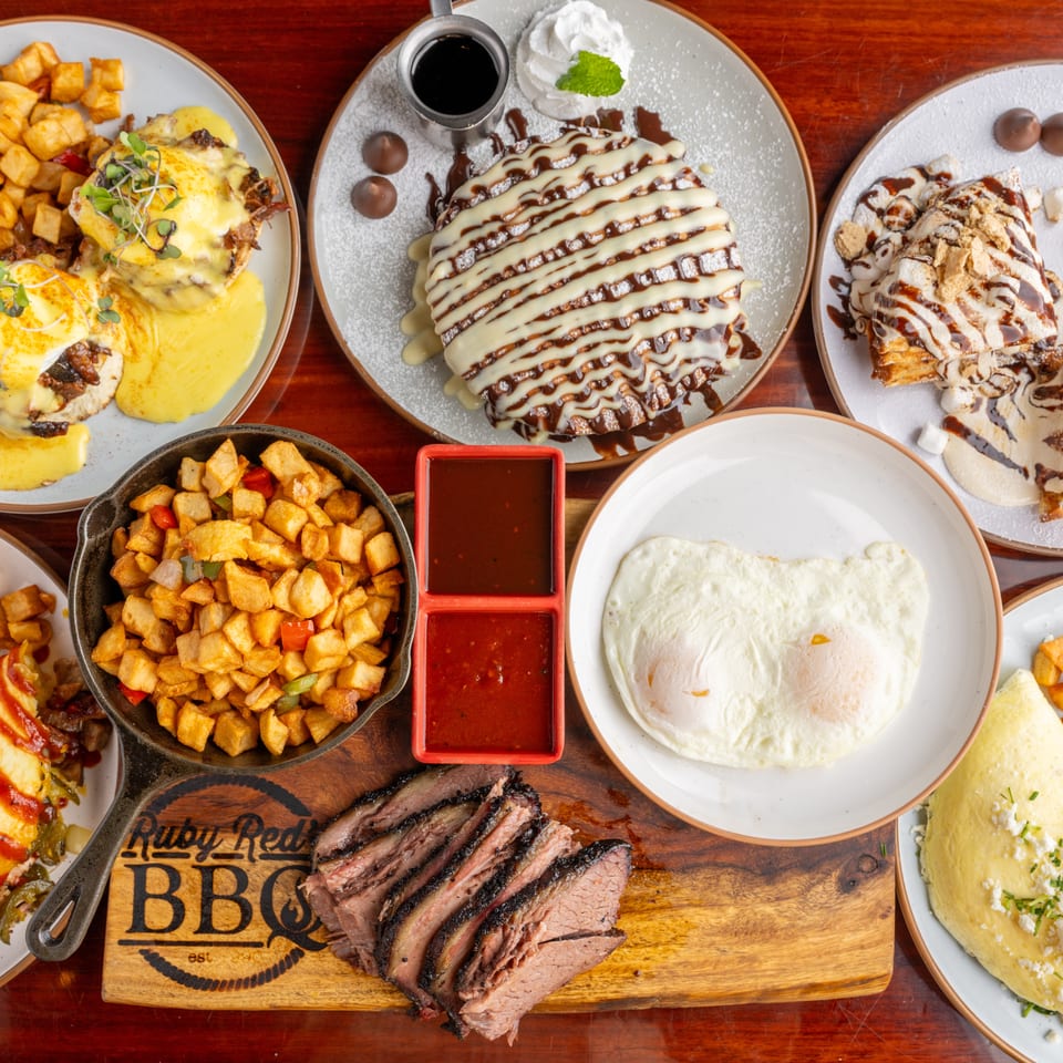 Overhead spread of barbecue brisket, sauced entrées, potatoes, desserts, dipping sauces, and sides on a wooden table.