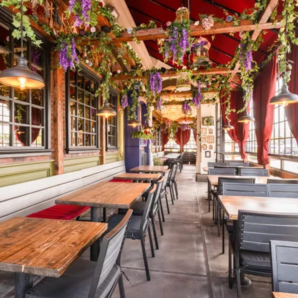 Bright restaurant dining room with wooden tables, black chairs, red curtains, and abundant hanging greenery and flowers.