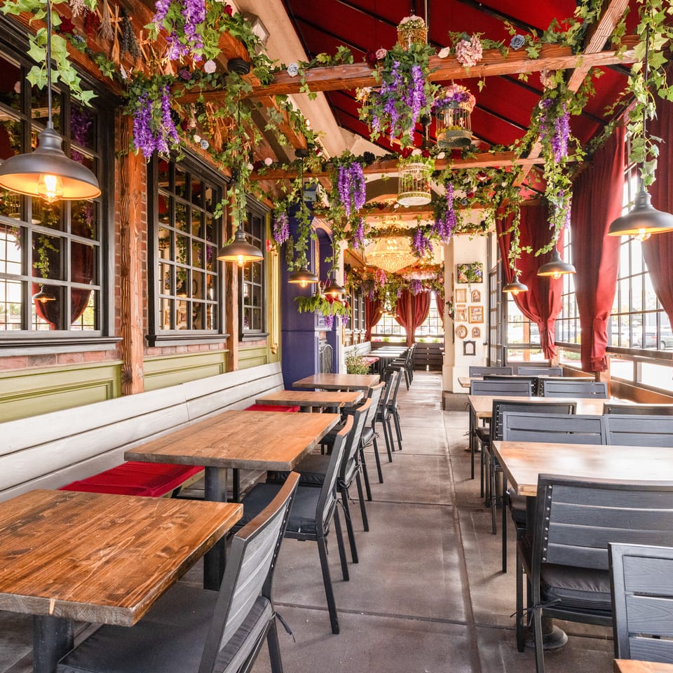 Restaurant dining room with wooden tables, black chairs, red curtains, pendant lighting, and abundant hanging flowers and greenery.