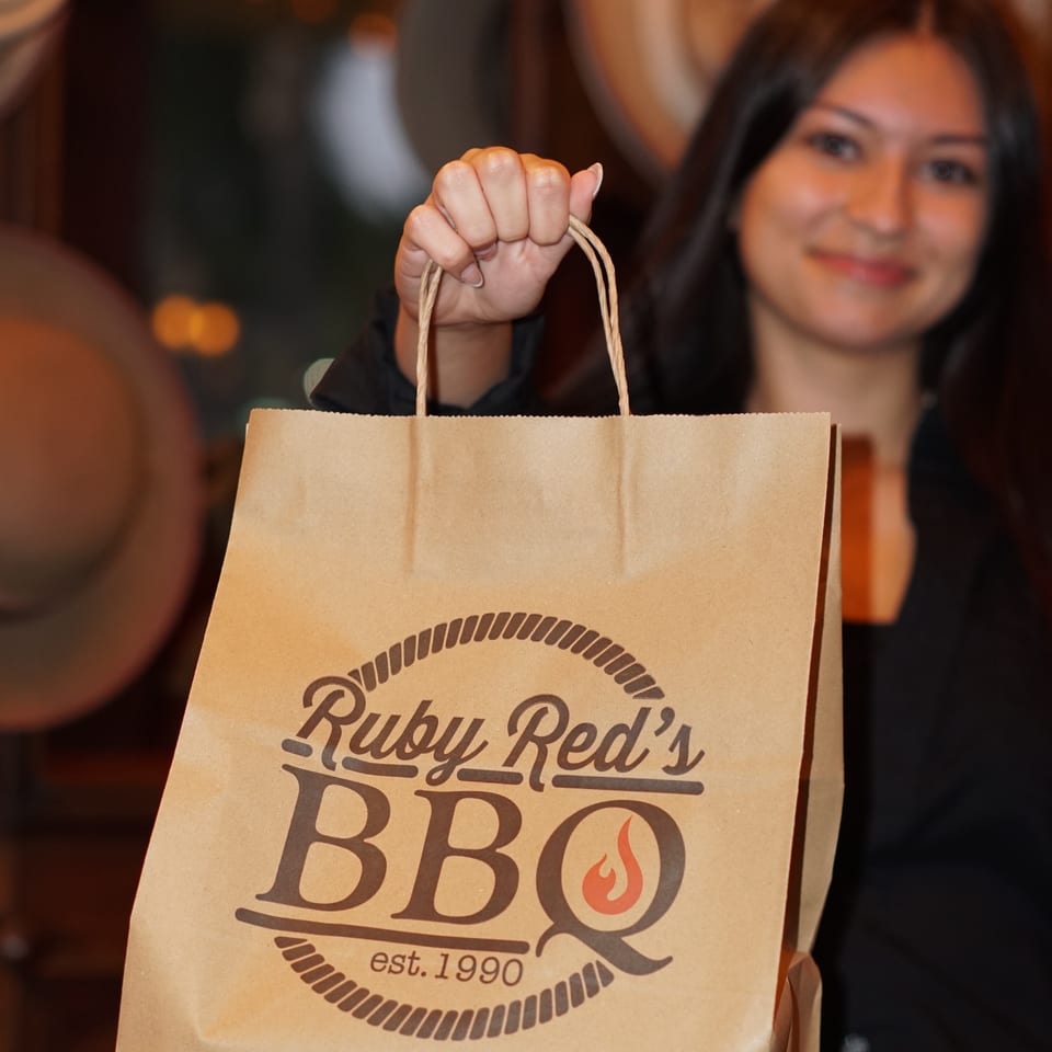 Smiling woman holds a Rudy Red’s BBQ paper bag inside a warmly lit restaurant.