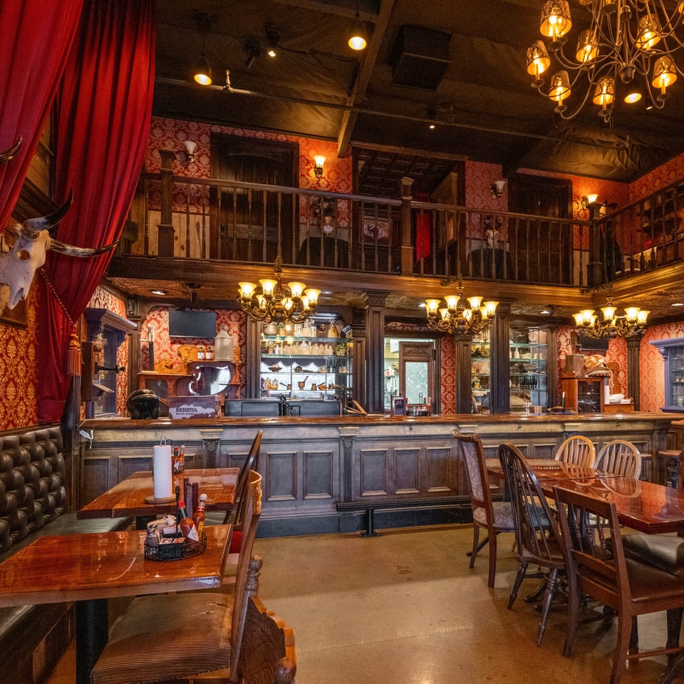Ornate restaurant interior with red velvet curtains, dark wood bar, chandeliers, patterned wallpaper, and dining tables.