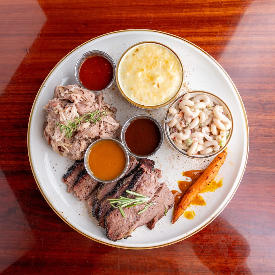 Overhead view of sliced roast meats with barbecue sauces, macaroni salad, cheesy casserole, and roasted carrots on a white plate.