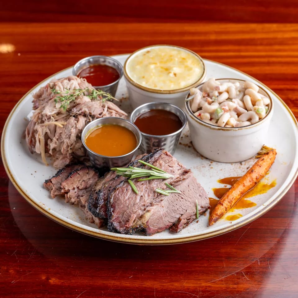 Platter of sliced roast beef, pulled pork, creamy macaroni, mashed potatoes, roasted carrot, and dipping sauces on a wooden table.