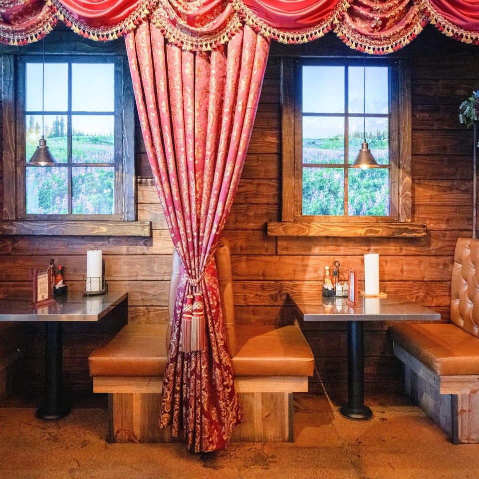 Rustic restaurant dining area with wood-paneled walls, leather booths, ornate red curtains, tables, and mountain meadow views through windows.