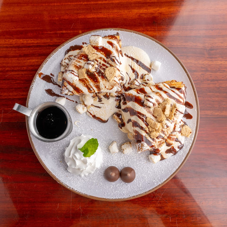 Dessert crepes topped with whipped cream, marshmallows, cookie crumbs, powdered sugar, and chocolate sauce on a plate.