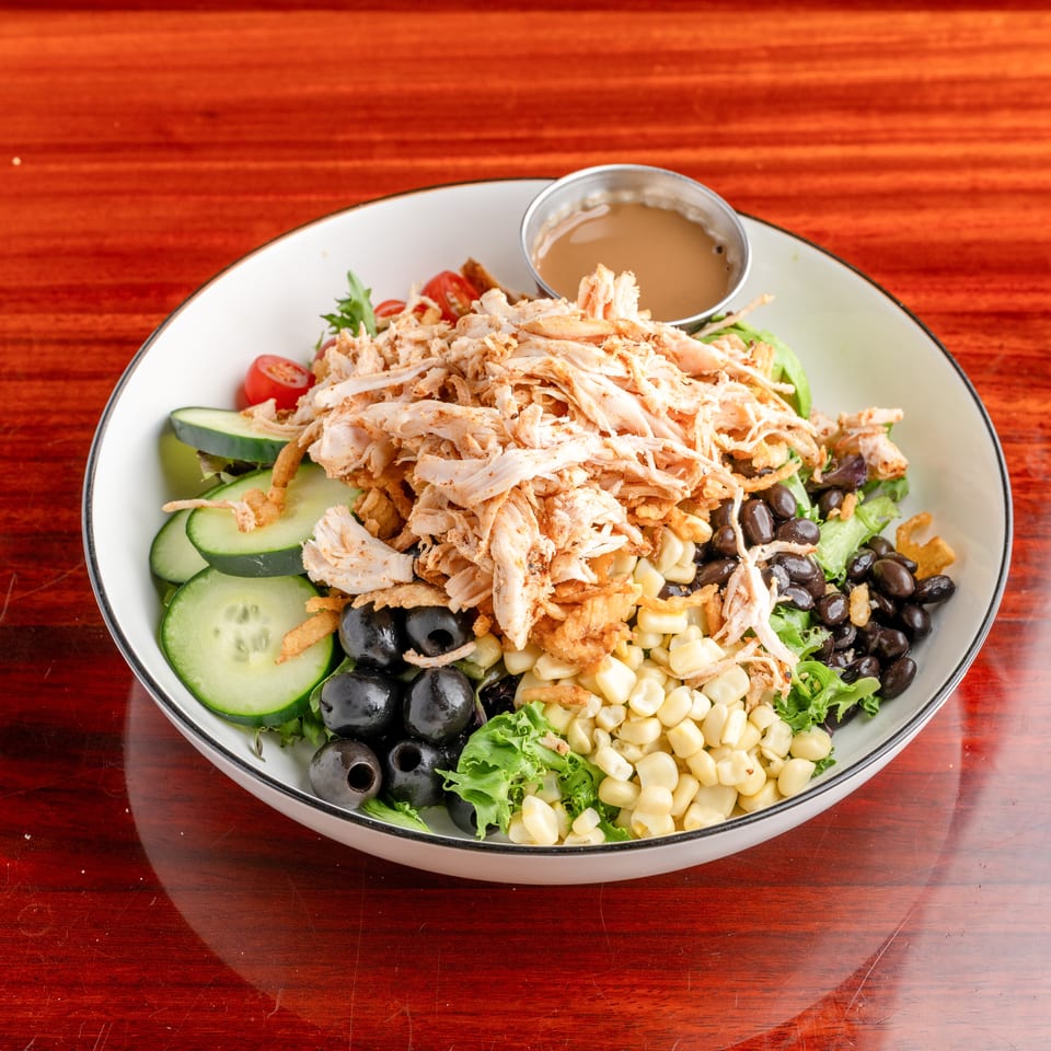 Large salad bowl topped with shredded chicken, cucumbers, corn, black olives, tomatoes, greens, crispy onions, and dressing.