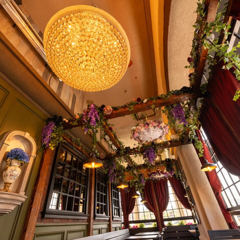 Elegant restaurant interior with a crystal chandelier, floral-covered wooden beams, burgundy curtains, and ornate decor.