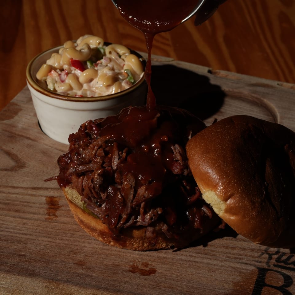 Barbecue sauce is poured over pulled pork sandwiches beside a cup of creamy macaroni salad on a wooden serving board.