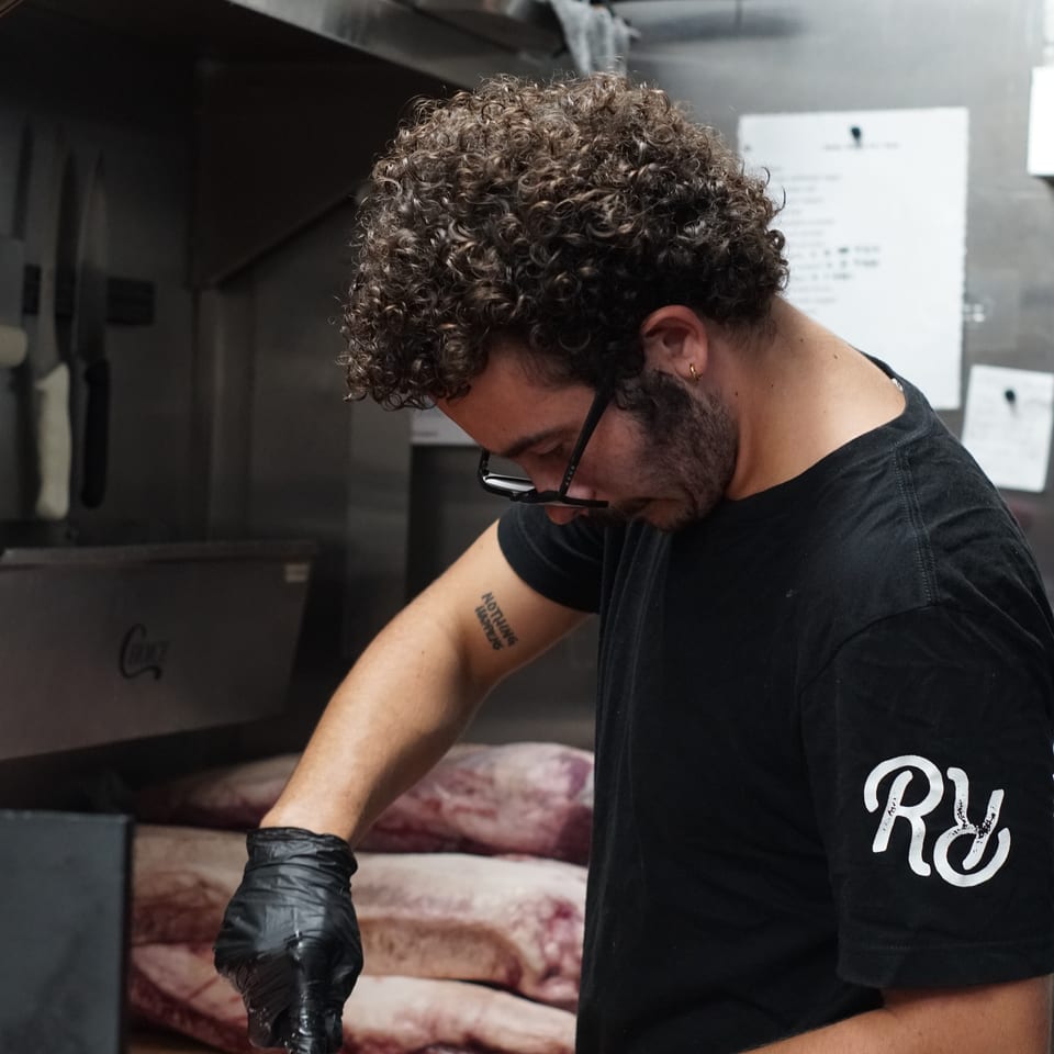 Restaurant worker in black gloves trims raw meat on a wooden cutting board in a commercial kitchen.