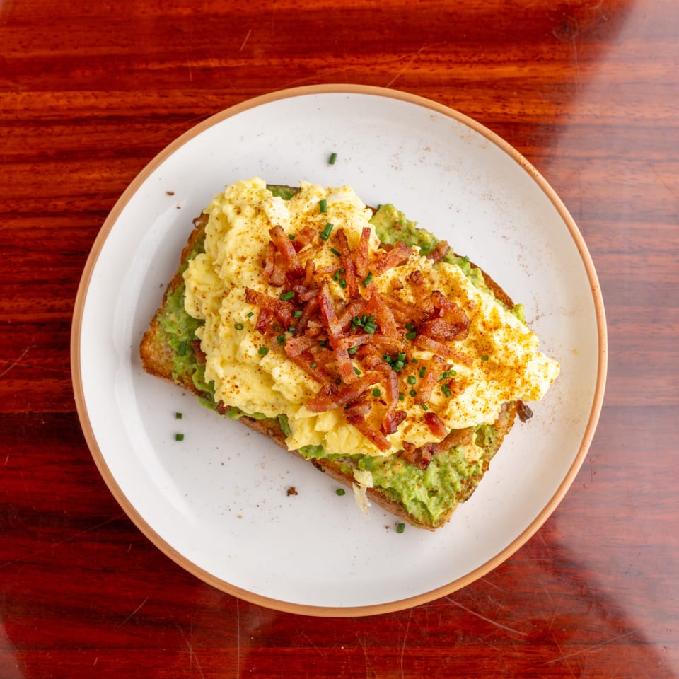 Avocado toast topped with scrambled eggs, crispy bacon, chives, and paprika on a white plate atop a wooden table.