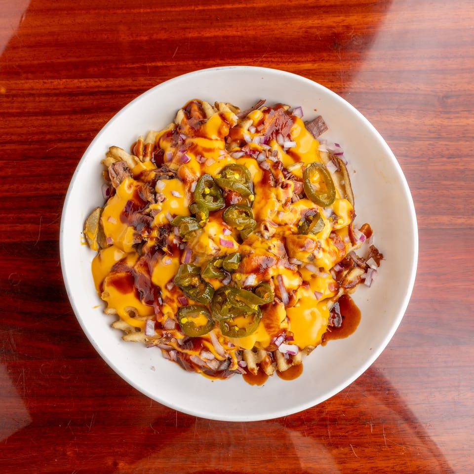 Loaded fries topped with melted cheese, barbecue sauce, pulled meat, jalapeños, and diced red onions in a white bowl.