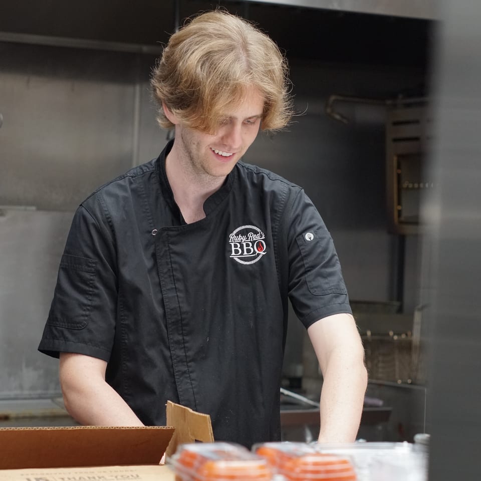 Smiling Ruby Red’s BBQ employee in a black uniform prepares food in a commercial kitchen beside containers of red sauce.