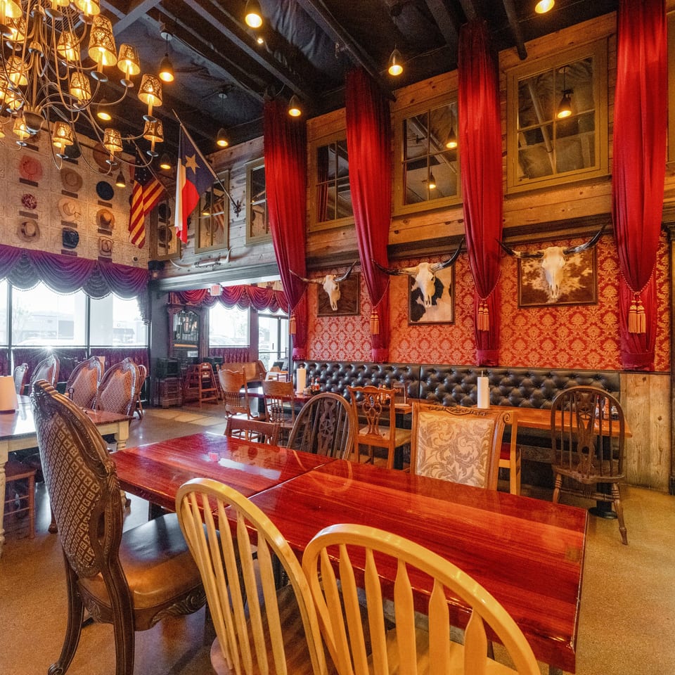 Ornate Western-themed restaurant dining room with red curtains, chandeliers, patterned walls, cow skulls, flags, and wooden tables.