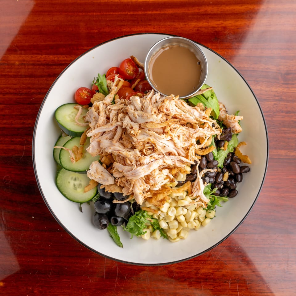 Overhead view of a salad topped with shredded chicken, cucumbers, tomatoes, avocado, black beans, corn, olives, and dressing.