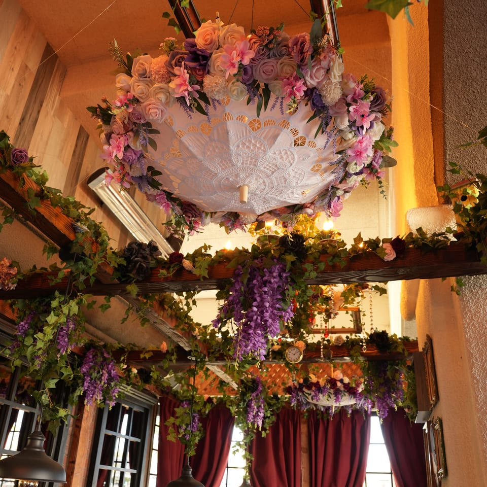 Ornate restaurant interior featuring a floral chandelier, cascading ivy, burgundy curtains, pendant lights, and diners by large windows.