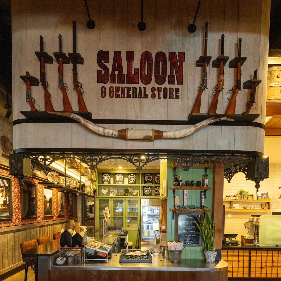Western-themed restaurant interior featuring a Saloon General Store sign, rifles, buffalo horns, rustic decor, and service counters.