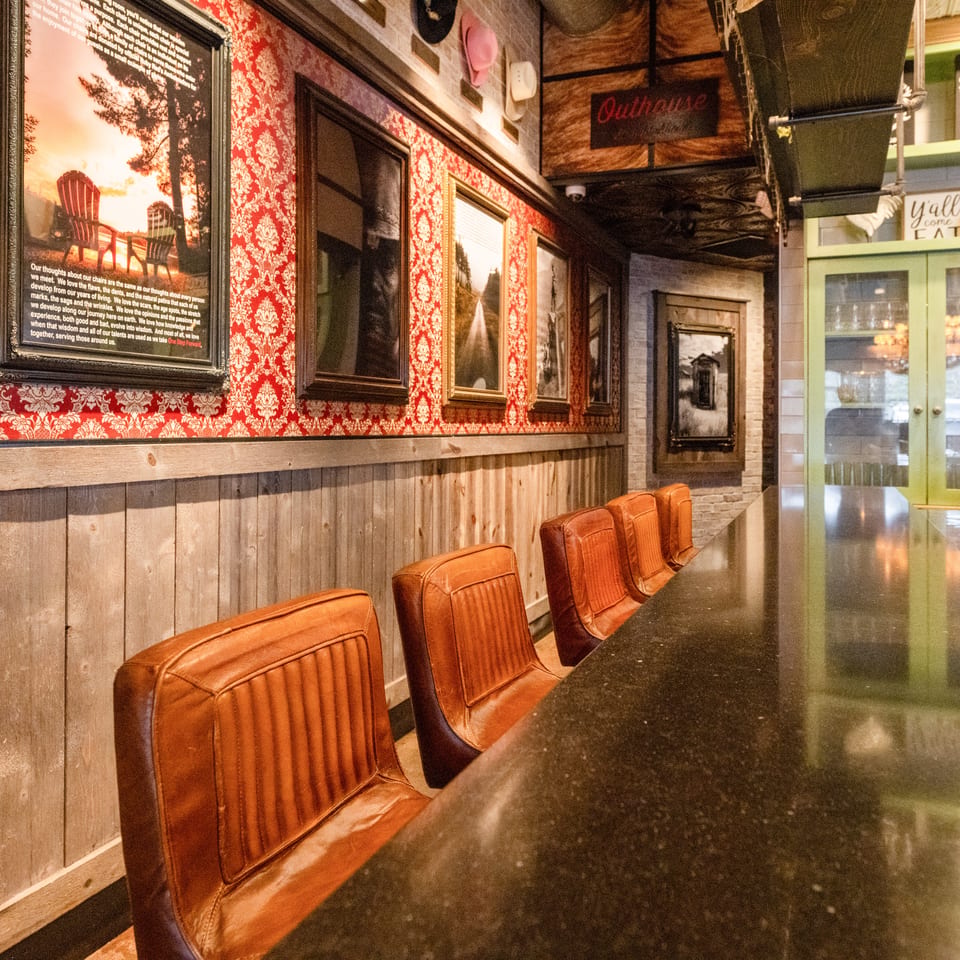 Restaurant dining area with leather chairs, a long dark countertop, rustic wood paneling, and ornate red patterned wallpaper.