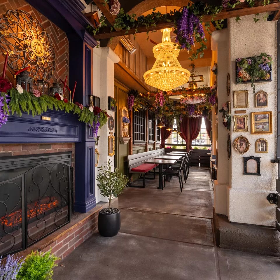 Eclectic restaurant interior with a glowing chandelier, flower garlands, ornate fireplace, framed art, and empty dining tables.