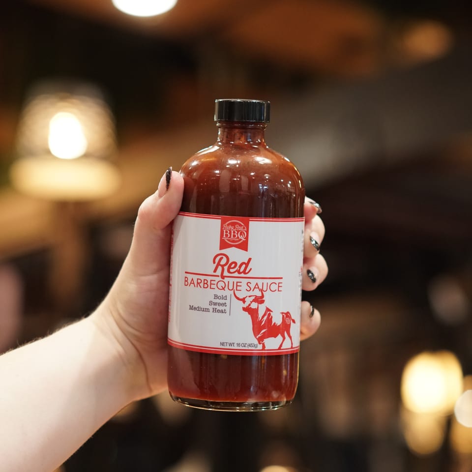 Hand holding a bottle of Ruby Red BBQ barbecue sauce against a warmly lit, blurred restaurant background.