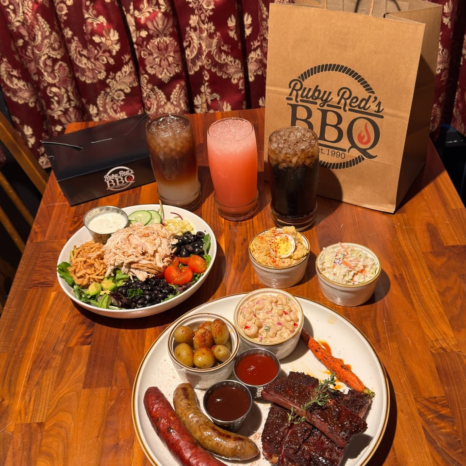 Ruby Red’s BBQ table spread with ribs, sausages, chicken salad, sides, drinks, and branded takeout bag.