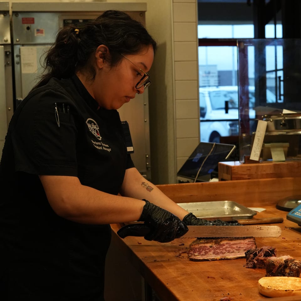 Restaurant worker wearing gloves prepares sliced barbecue meat on a wooden cutting board behind the counter.