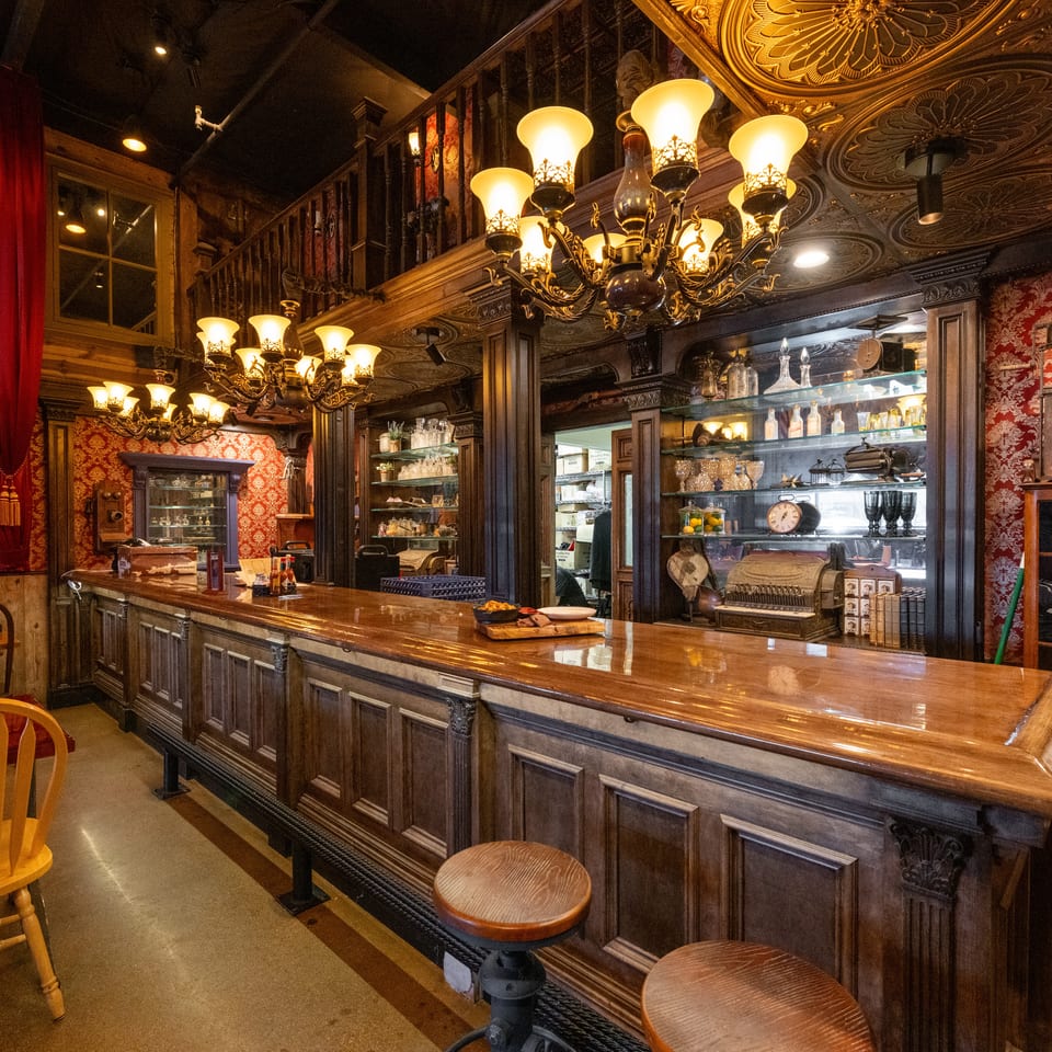 Ornate restaurant bar with dark wood paneling, red damask wallpaper, mirrored shelves, vintage chandeliers, and stools.