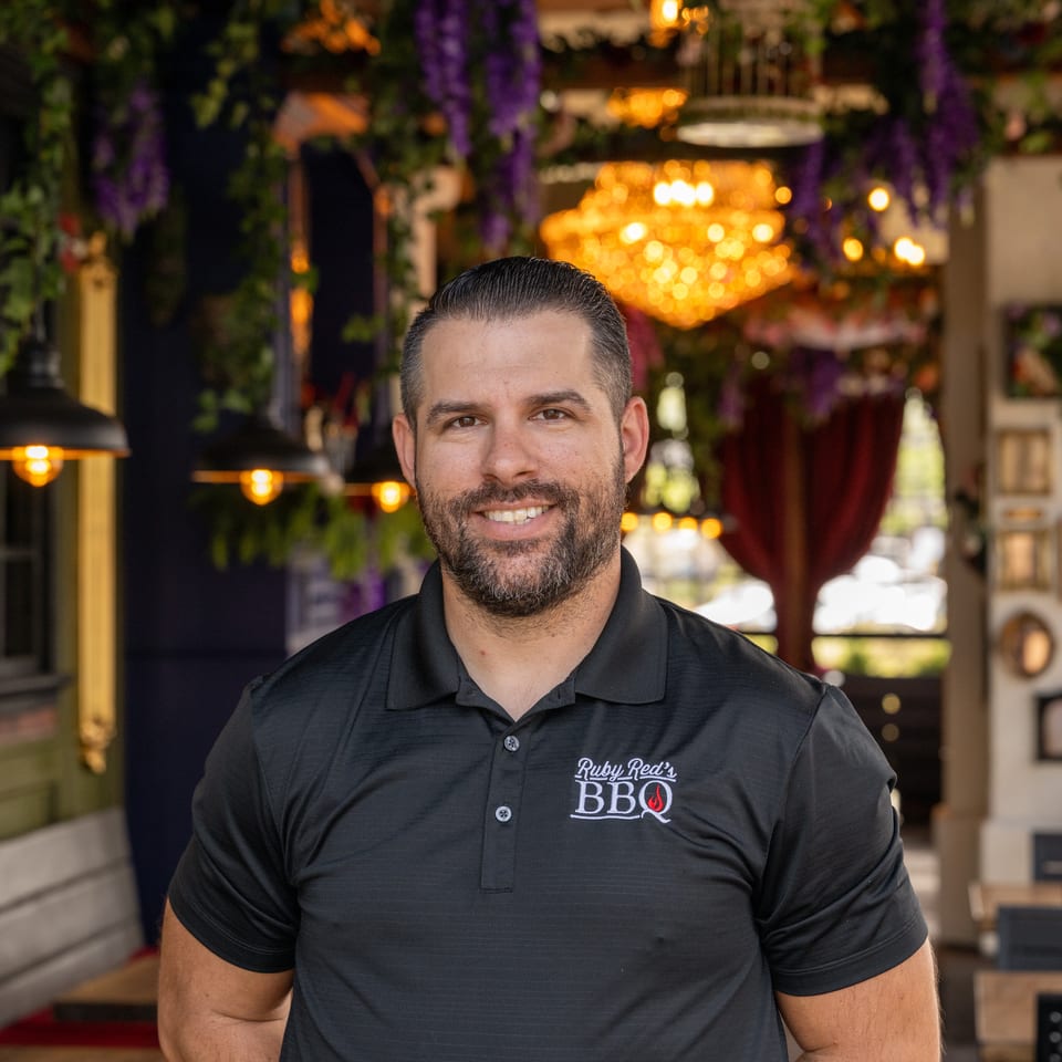 Smiling man wearing a Ruby Red’s BBQ polo stands inside a warmly lit, plant-filled restaurant with chandeliers.