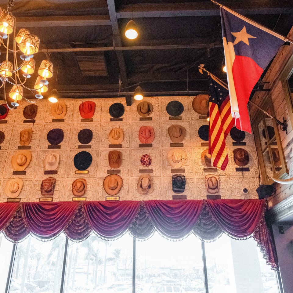 Restaurant interior featuring a wall-mounted collection of cowboy hats, ornate lighting, draped curtains, and colorful flags.