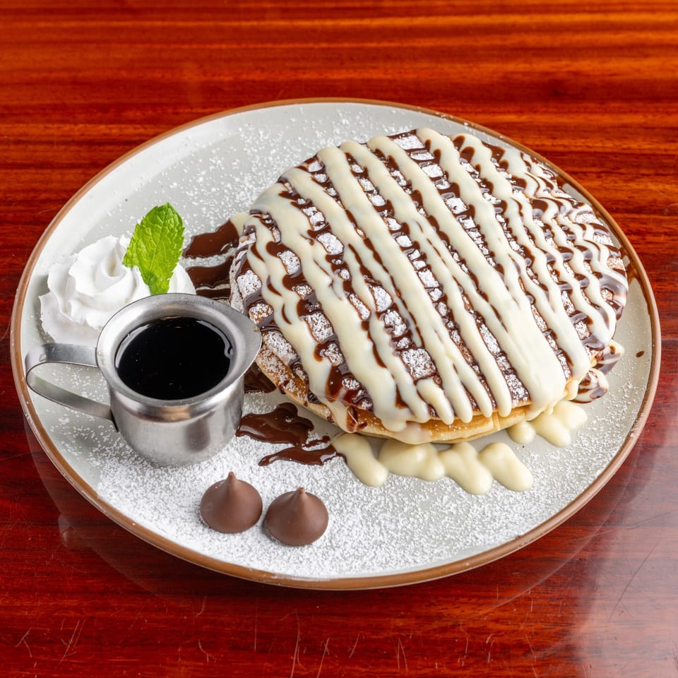 Chocolate-drizzled pancakes topped with white sauce and powdered sugar, served with whipped cream, mint, chocolate sauce, and truffles.