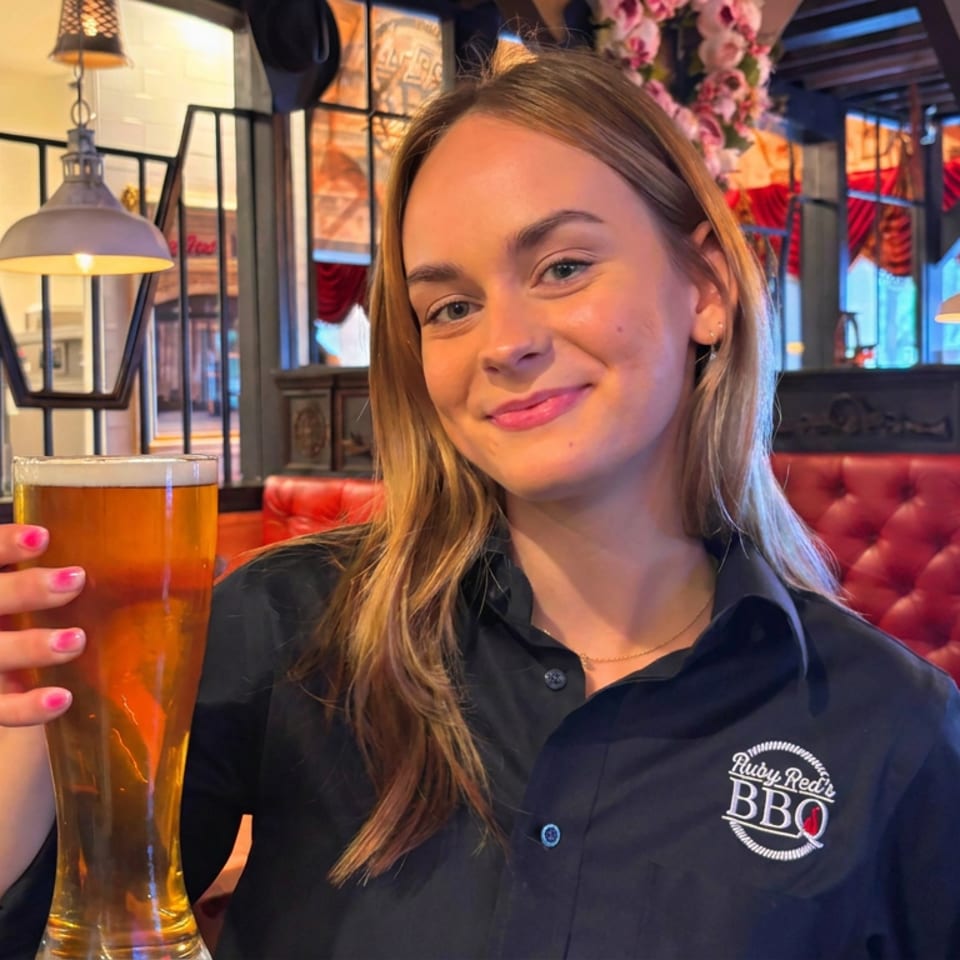 Smiling restaurant server holds a pint of amber beer inside a warmly lit barbecue restaurant with red booths and floral décor.